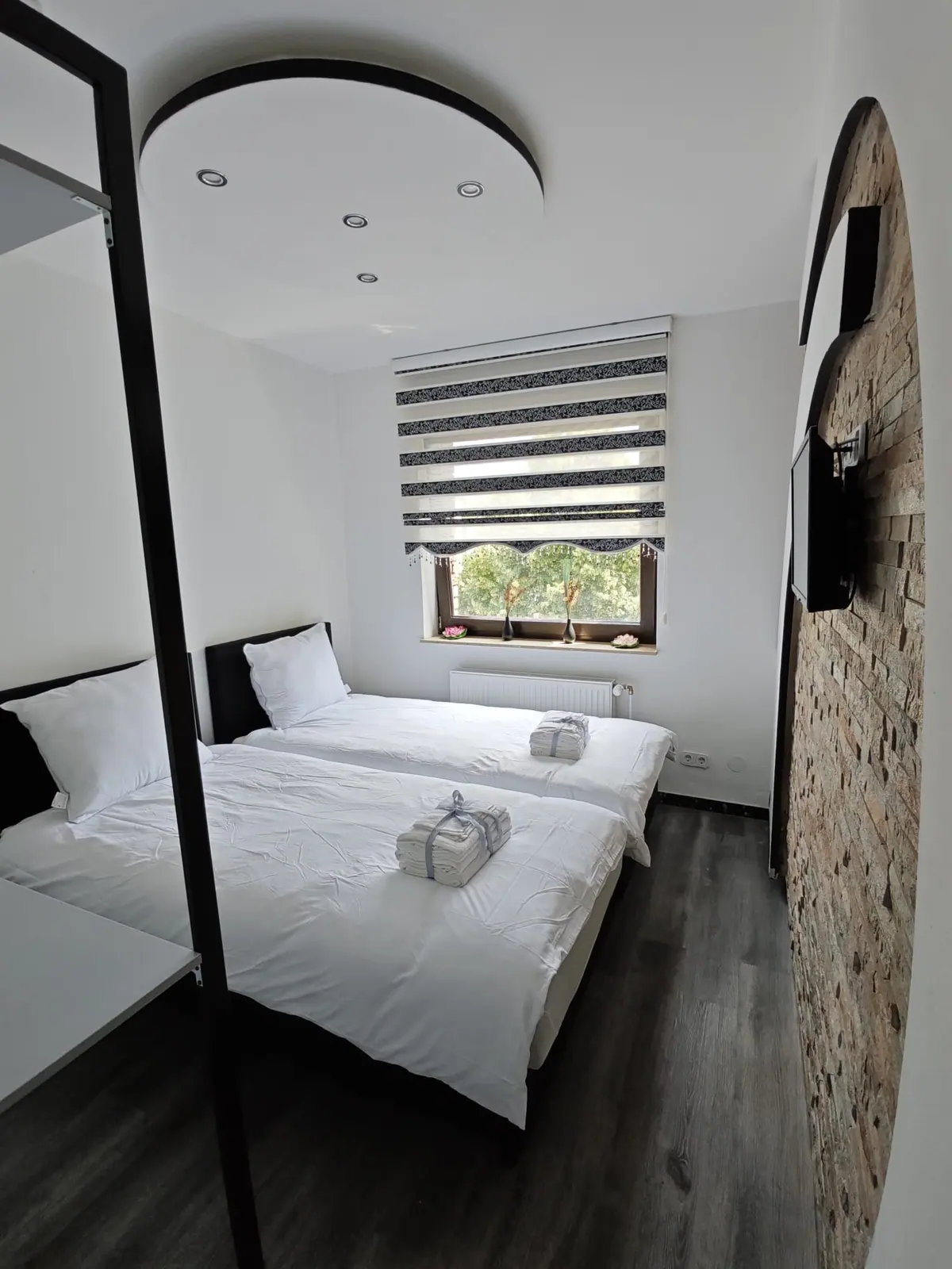 Zweibettzimmer mit hellem Sandsteinbogen, Wandfernseher und rundem Deckenausschnitt mit Spots