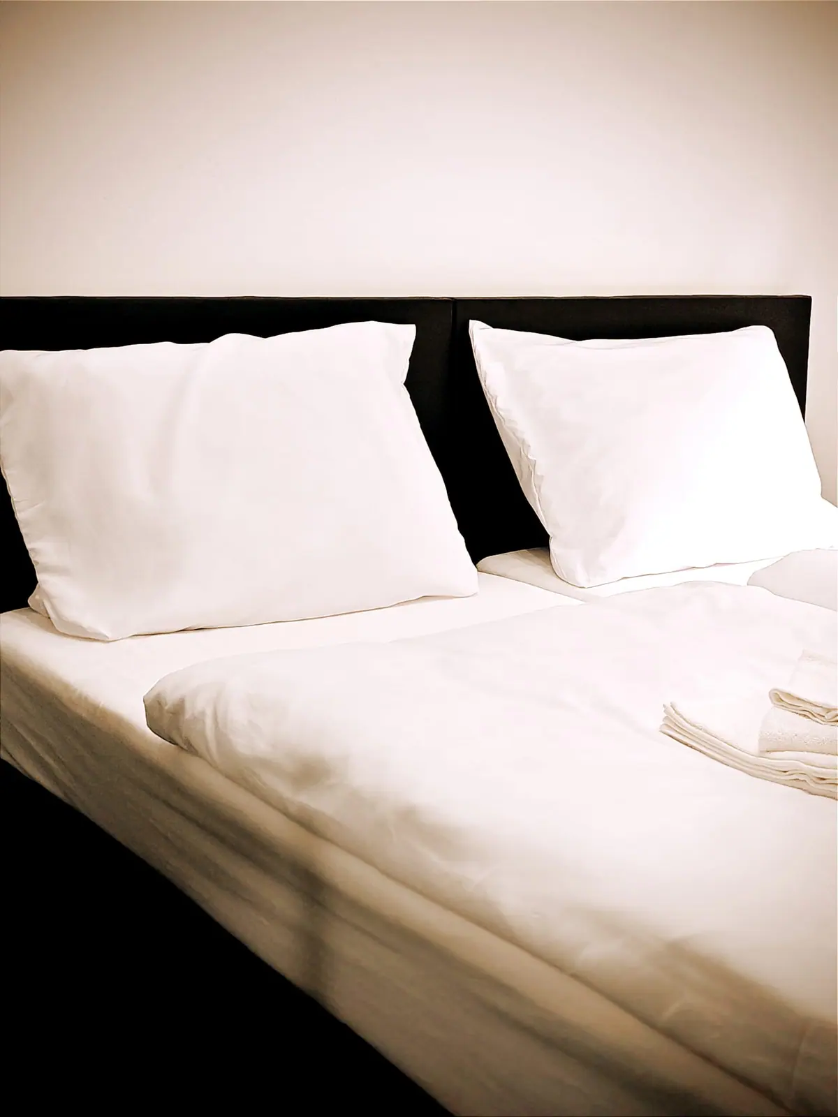 Doppelbett mit zwei weißen Kissen vor schwarzem Kopfteil und frischen Handtüchern