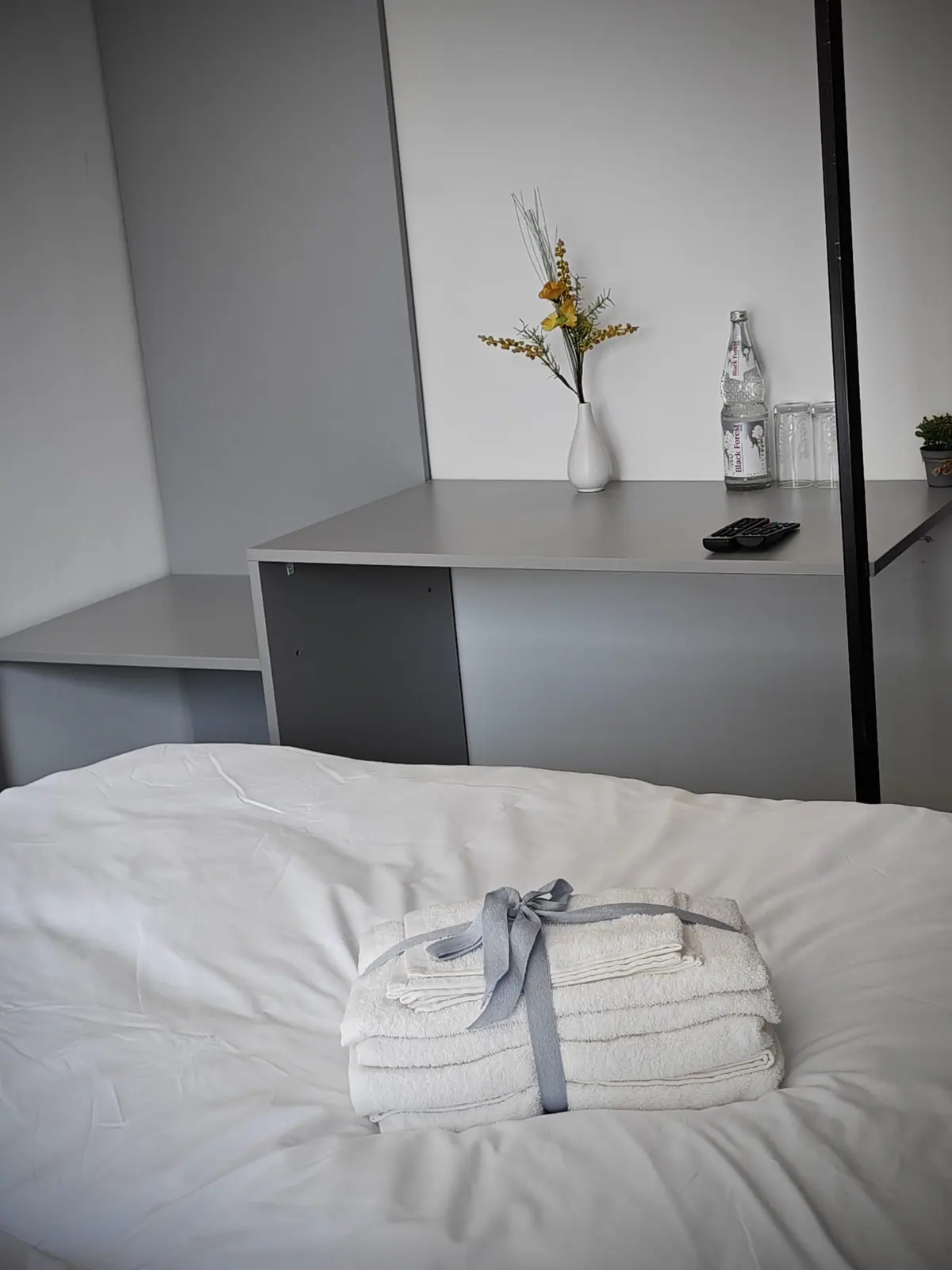Einzelzimmer mit Schreibtisch, Wasserflasche, Vase und Handtüchern auf dem Bett